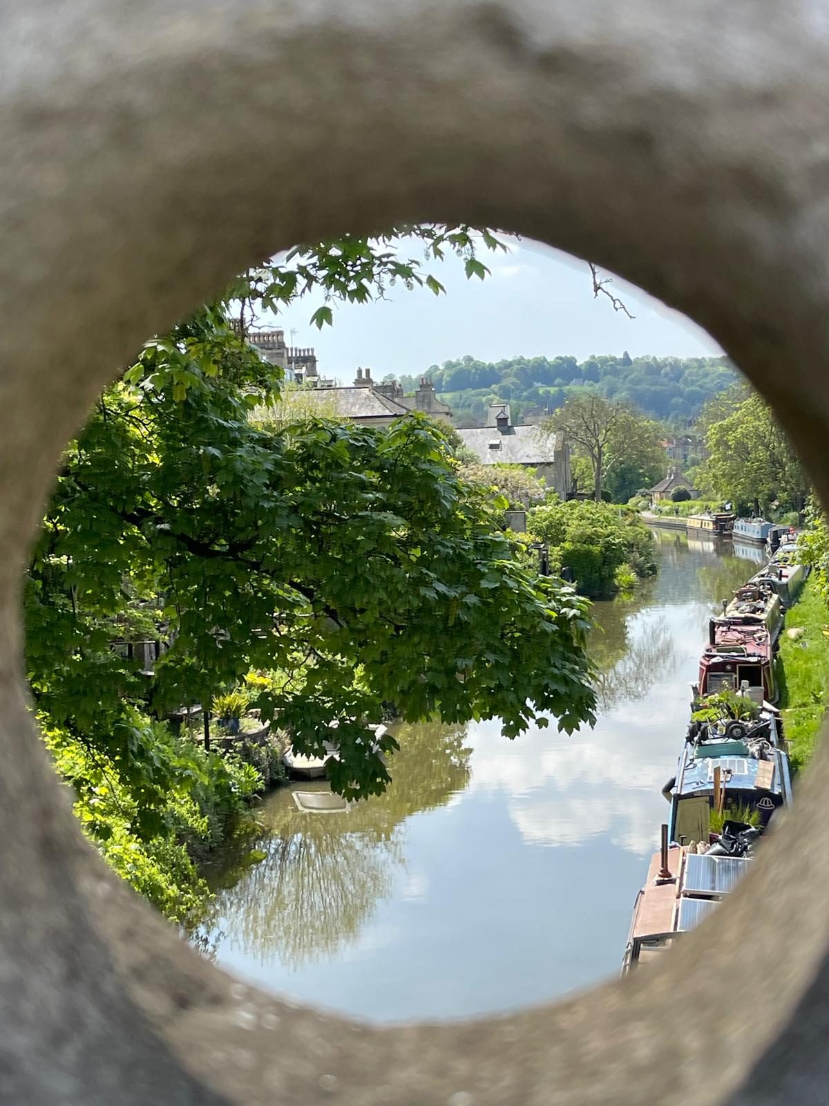 A view down an English canal, framed through a circular stone aperture in a bridge. A line of moored narrowboats stretches into the distance, with overhanging trees on the opposite bank and hills beyond.